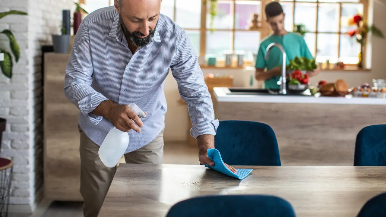 Man cleaning dining table while someone cooks nearby..