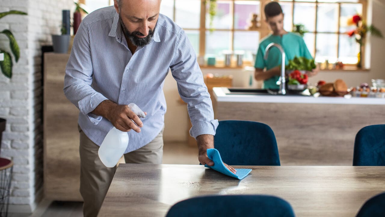 Man cleaning dining table while someone cooks nearby..